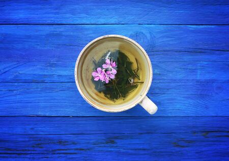 Natural herbal tea with purple fresh flowers and leaves of medical fireweed plant n ceramic cup on blue wooden boards outdoors.の写真素材