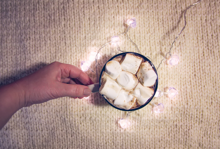 Woman's hand holding ceramic mug with hot fresh cocoa drink with marshmallows. Decorative garland lights on cozy knitted background.の写真素材