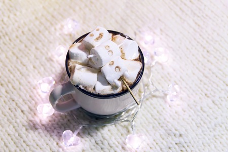 Ceramic mug with hot fresh cocoa drink and melted handmade funny marshmallow snowman. Decorative garland lights on cozy knitted background.の写真素材