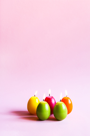 Traditional Easter decor. Group of bright burning paraffin candles in the shape of colorful eggs on soft pink background.の写真素材