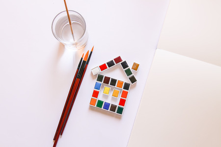 Watercolor paints in the shape of a funny house, water in glass, brushes and paper on white background. Painting tools. Flat lay, top viewの写真素材