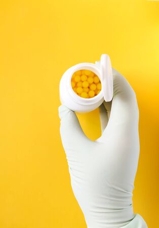 Hand in rubber glove holds vitamin C pills in a white plastic storage on bright yellowの写真素材