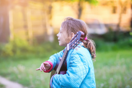 Little girl playing guitar and singing outdoors on green meadow at spring. Happy child on rural yard.の写真素材