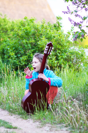 Little girl playing guitar and singing outdoors on green meadow at spring. Happy child on rural yard.の写真素材
