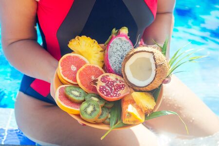 orange, coconut, pomegranate, dragonfruit, pineapple on a plate in the hands of a woman in a swimsuit against the background of the swimmingpool. detox.の写真素材