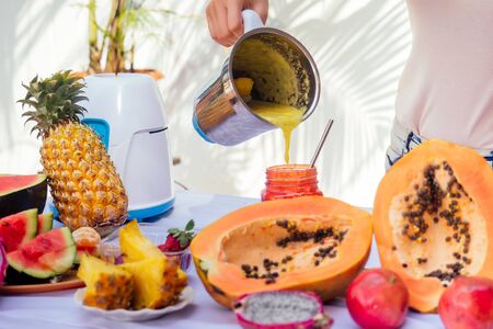Lifestyle blonde woman blending blender juice glass jars with smoothies and straw in the summer balcony.tropical exotic fruits on the table.cleansing the body of toxins idea.summertime season.の写真素材