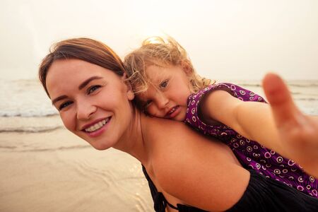 Happy mother and daughter taking selfie photo with smartphone on the Sunset beach. woman having fun...
