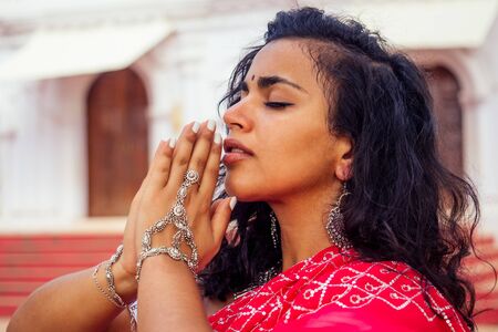 Young Indian woman in traditional sari red dress praying in a hindu temple goa india Hinduism.girl performing namaste gesture catholicism Delhi Street holi festival.om yoga meditation female modelの写真素材