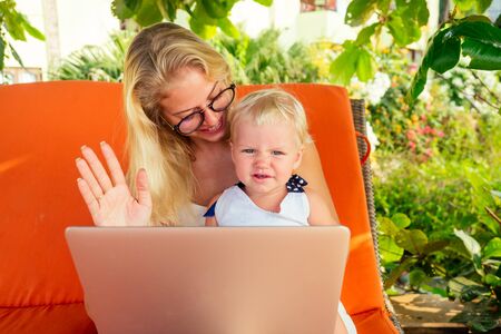 beautiful blonde woman and little girl are looking into the screen laptop and waving their hands hi sea vacation.young mother,fair-haired one year old daughter talking communication relatives onlineの写真素材
