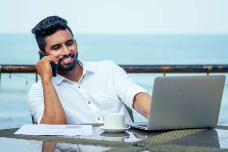 handsome and successful indian man in a stylish well-dressed freelancer work laptop on the beach.freelance and remote work.businessman student in a summer cafe cup of tea on the shore of india oceanの写真素材