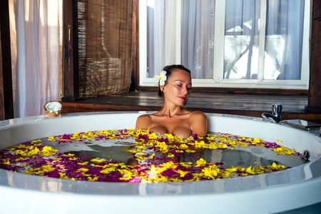 Beautiful girl brunette with plumeria in her hair in tub with rose petal and tropical flowers. Body...