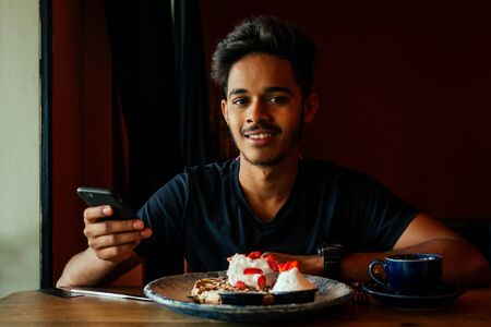 young male drinking latte at coffee bar pancakes at cafe and holding smartphoneの写真素材