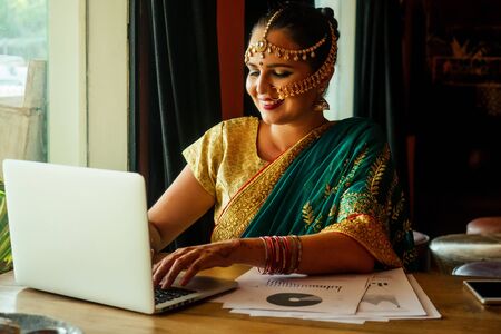 beautiful looking indian young girl in green sari working on laptop and using credit ATM card payment online, sitting in cafe remote work and freelanceの写真素材
