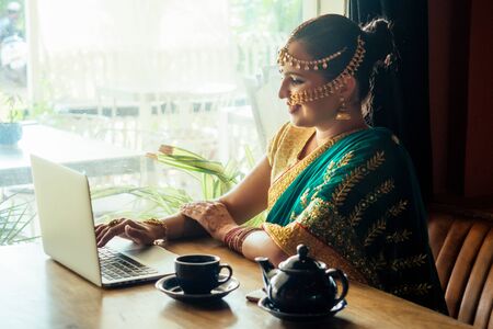 pretty indian businesswoman in green traditional sari using laptop in office next to window. remote work and freelanceの写真素材