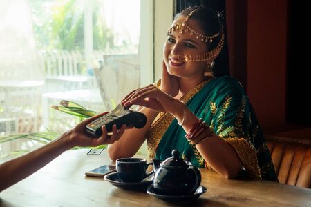 Young indian businesswoman in green traditional sari on a coffee break. Using payment terminal...
