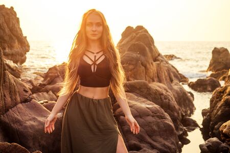 fashion hippie model portrait outdoors boho style young flower children woman with headdress made of feathers in a brown long skirt and black top dress,stylish shoes boots on stone Arambol Goa beachの写真素材