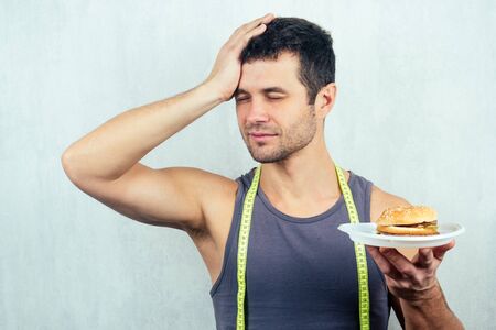 portrait of a young beautiful brunette man with a measuring tape eating a high-calorie big burger on a plate with a fork. concept of diet, unhealthy and healthy eatingの写真素材