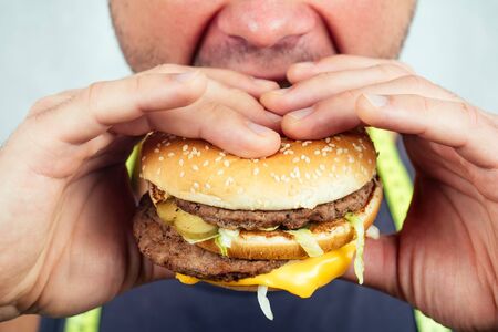 portrait of a young beautiful man eats bites a high-calorie big burger in his hands. concept of diet, unhealthy and healthy eatingの写真素材