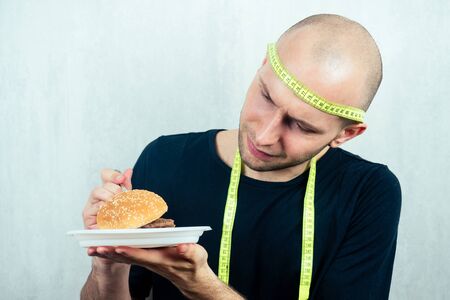 portrait of a young handsome bald man with a measuring tape eating a high-calorie big burger on a plate with a fork. concept of diet, unhealthy and healthy eatingの写真素材