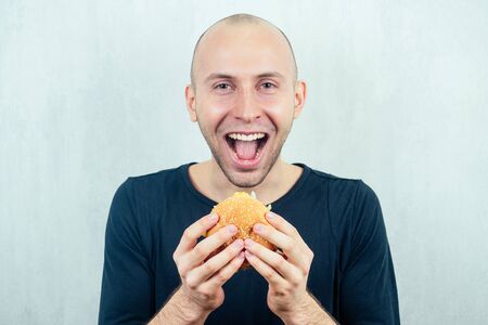 Portrait of a young handsome bald man holding a high-calorie big burger in his hands. concept of diet, unhealthy and healthy eatingの写真素材