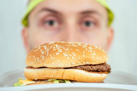 portrait of a young handsome bald hunger man with a measuring tape looking at a high-calorie big burger on a plate. concept of diet, unhealthy and healthy eatingの写真素材
