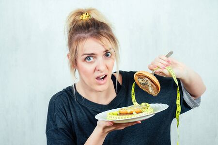 portrait of a hungry, young and beautiful blonde woman holding a burger cheeseburger on a plate in a...