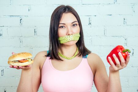 portrait of a beautiful and young woman with a knotted tape measuring tape making a choice between a calorie big burger and pepper in the gym. concept of a quick bad snack and a vegetable dietの写真素材