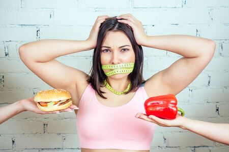portrait of a beautiful and young woman with a knotted tape measuring tape making a choice between a calorie big burger and pepper in the gym. concept of a quick bad snack and a vegetable dietの写真素材