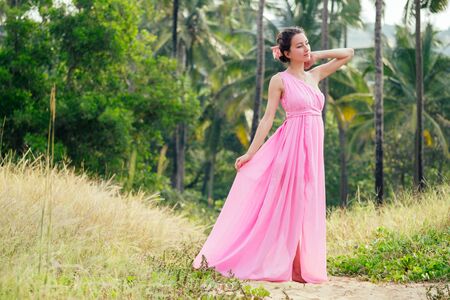 young and beautiful woman posing in an elegant pink long chic luxury dress against a background of palm trees. concept of chic vacation in a tropical resortの写真素材