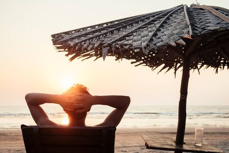 the man is resting on a sun lounger overlooking the sea and the sunset on the beachの写真素材