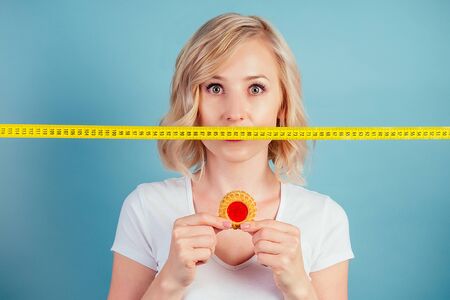attractive sad blonde woman with make-up and lips color fuchsia hold a high-calorie cookie biscuit iwith measuring tape n the studio on a blue background . diet and rejection of flour productsの写真素材