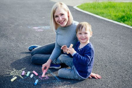 beautiful blonde mother and her cute baby son painted with colored chalk crayons on the asphalt in the park on a background of green grassの写真素材