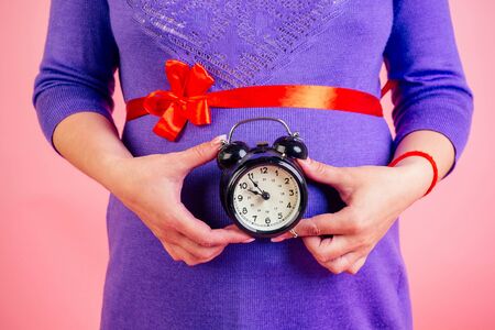 close-up hands of pregnant woman in a purple dress and a red satin bow on belly holding a alarm clock in studio on a pink background . time for birth conceptの写真素材