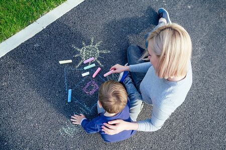 beautiful blonde mother and her cute baby son painted with colored chalk crayons on the asphalt in the park on a background of green grass view from aboveの写真素材