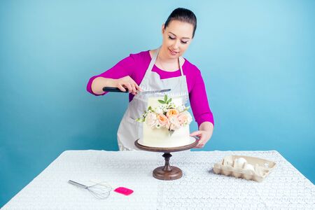 confectioner female person pastry-cook decorate appetizing creamy white two-tiered wedding cake decorated with fresh flowers on a table with a lace tablecloth in studio on a blue backgroundの写真素材