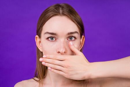 portrait of a frightened woman model holding close mouth shut up hand on purple background in studio