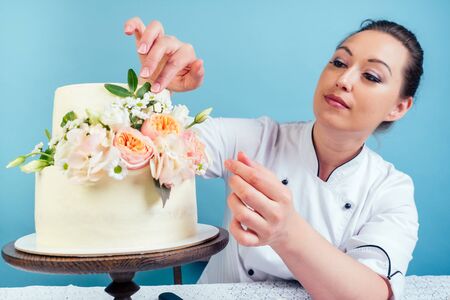 confectioner woman pastry-cook in confectioners jacket decorate appetizing creamy white two-tiered wedding cake with fresh flowers on a table with a lace tablecloth in studio on a blue backgroundの写真素材
