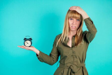 sad blonde woman keeps an alarm in hand in studio on blue background. concept of being late for work