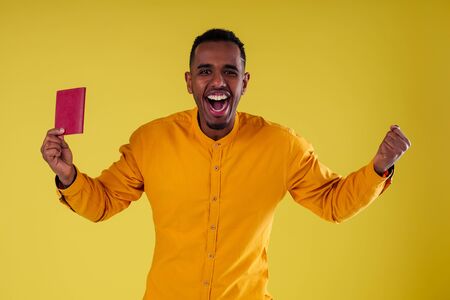 handsome happy young african man Kenya Africa tourist isolated yellow wall background holding tickets with passport and coctail glass trip around the worldの写真素材