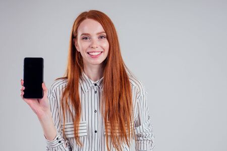 Cheerful redheared woman businesswomen in a striped shirt showing blank smartphone screen white background studioの写真素材