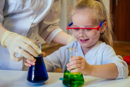 surprised and amazed clever blonde school girl chemist in eyeglasses and white medical gown making...