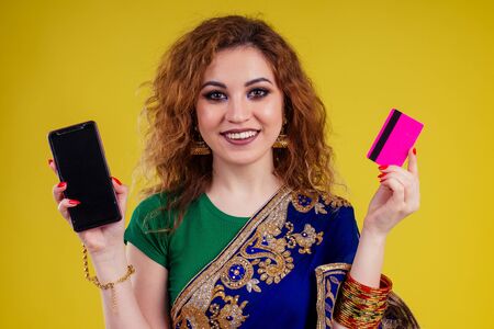 happy beautiful indian female person in traditional dress holding shop credit card and smartphone...