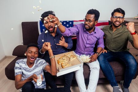 african american males eating pizza and popcorn , cheering and smiling while watching TV match at homeの写真素材