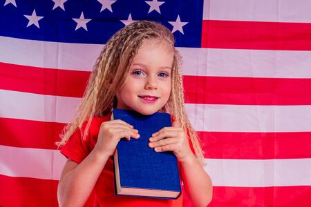 Little Blue eyes blonde charming girl holding an encyclopedia book alphabet foreign language against american flag backgroundの写真素材