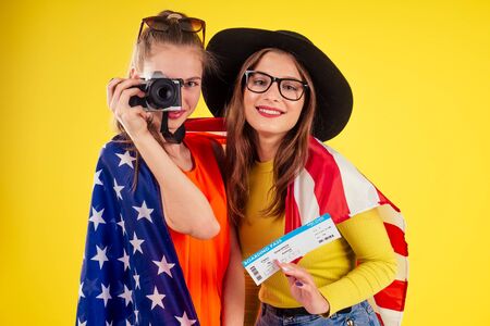 group of three laughing girls holding passports and tickets with photo camera wrapped in american flag spring party summer style yellow background studio, visa and independence day usaの写真素材