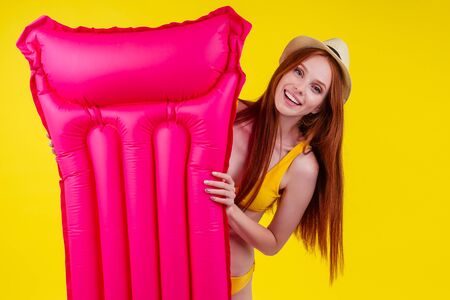 happy redhaired ginger woman ready to pool party,with pink mattress in studio wall yellow background wearing straw hatの写真素材