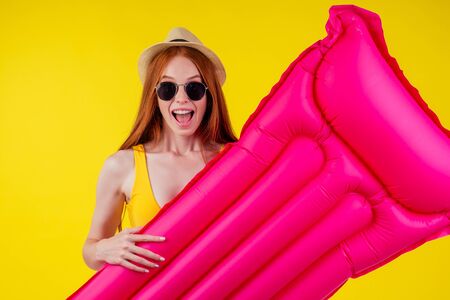 happy redhaired ginger woman ready to pool party,with pink mattress in studio wall yellow background wearing straw hatの写真素材