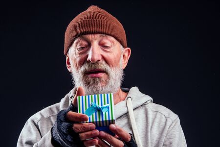 smilley portrait of old senior male tramp wearing warm brown hat and leaky gloves,holding blue gift box in studio black backgroundの写真素材