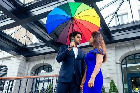 indian woman in long evening blue dress spinding romantic time together with lover handsome boyfriend under multi-colored rainbow umbrella europe urban downtown cityの写真素材
