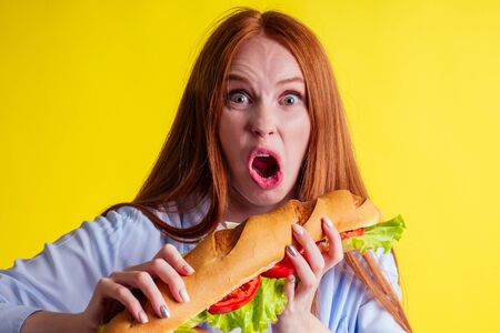 hungry redhaired ginger woman holding huge sandwich with pepper and lettuce salad between bun in studio yellow backgroundの写真素材